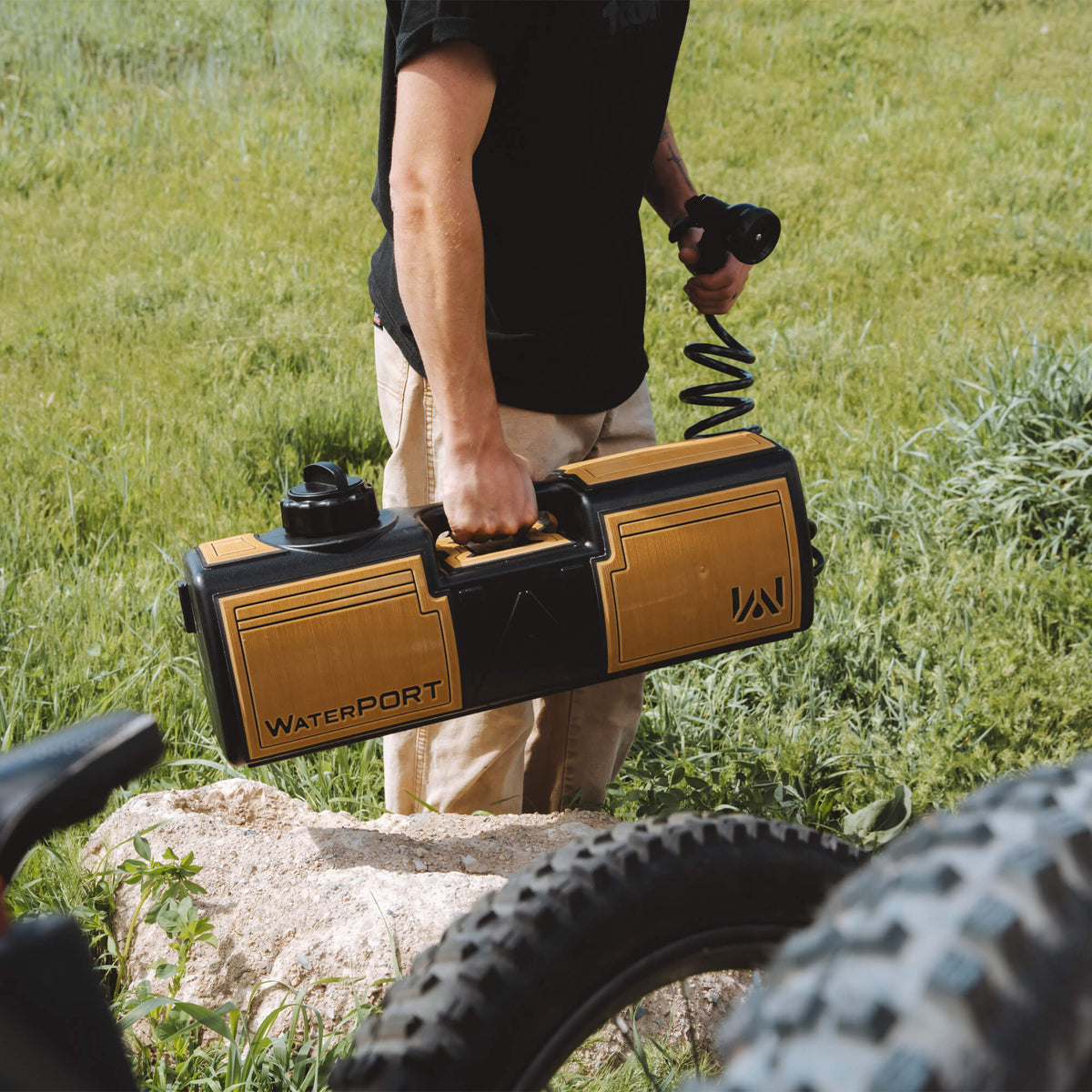An individual outdoors holds the EVA Wrap - 4 Gallon portable water tank with a sprayer hose, ideal for overland enthusiasts. In the background are green grass and a rock, while a bicycle tire appears in the foreground.