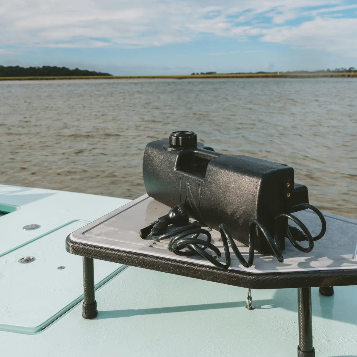 A black electronic device with coiled cords sits on a table next to a DayTank - 4 Gallon aboard a boat, with a river and grassy shoreline visible under a partly cloudy sky in the background.