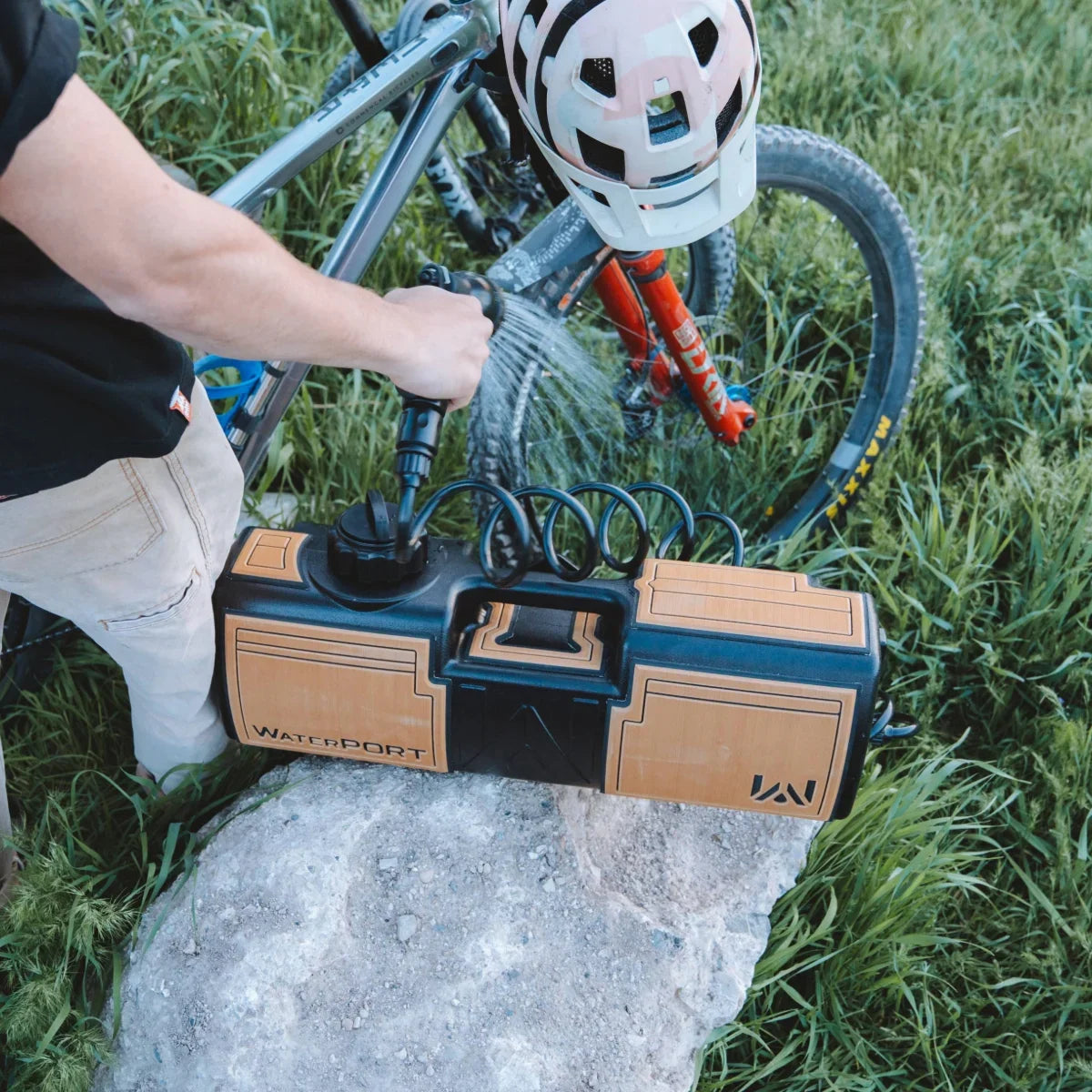 A person uses a handheld WaterPort spray nozzle to clean a mountain bike outdoors.