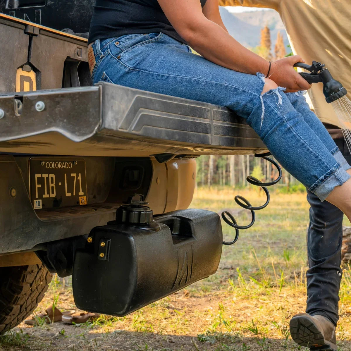 A person sits on a truck tailgate while using a coiled hose connected to a black water tank mounted under the vehicles bumper.