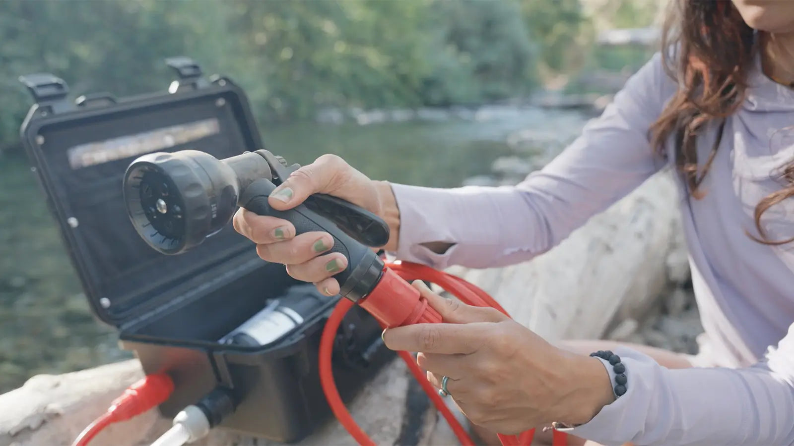 Person holding a spray nozzle and hose with a  HydroSpray sitting on a log in a natural setting beside a body of water