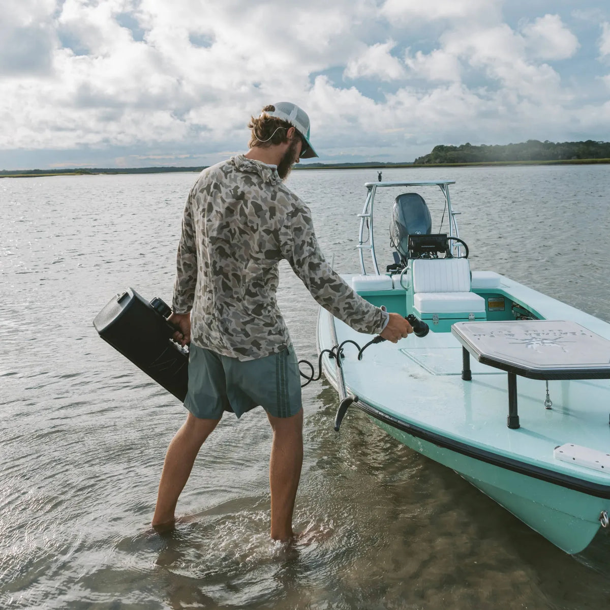 A man in a camo shirt, shorts, and cap stands in shallow water beside a blue boat, holding a DayTank - 4 Gallon portable pressurized water tank. Calm water and cloudy sky stretch out behind him.
