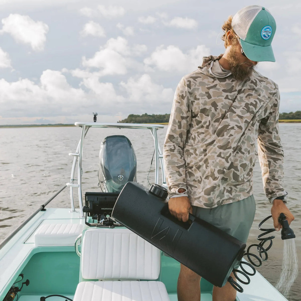 A man in a camo hoodie and teal cap stands on a light blue boat, holding a DayTank - 4 Gallon with a hose. The boat floats on calm water under a partly cloudy sky.