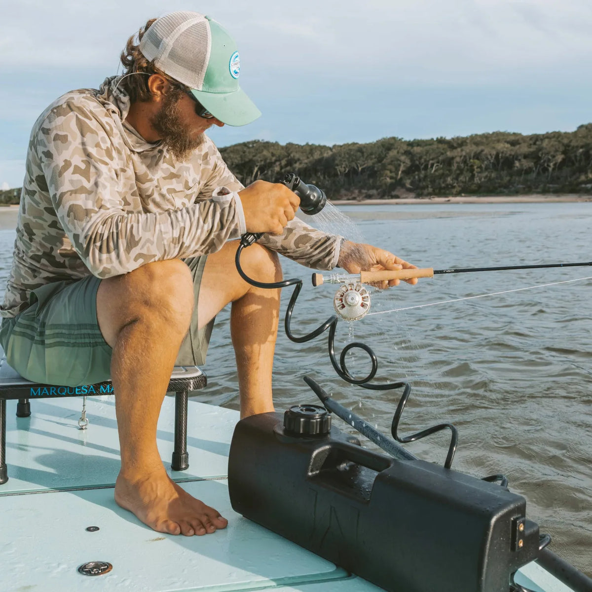 A man in a cap and camo shirt sits on a boat, rinsing a fishing rod with a hose from a DayTank - 4 Gallon portable pressurized water tank. Trees and shoreline are visible in the background.