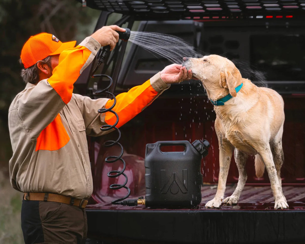 Man using a WaterPORT 2-gallon GoSpout spray nozzle to rinse a muddy yellow Labrador on a truck tailgate.