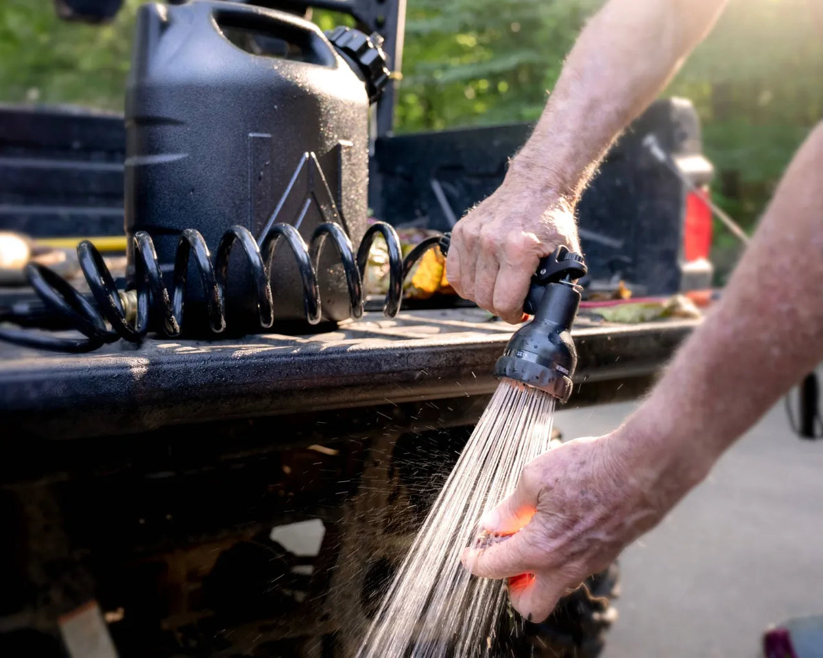 Hands rinsed under a steady shower spray from the WaterPORT 2-gallon GoSpout nozzle beside a pickup truck.