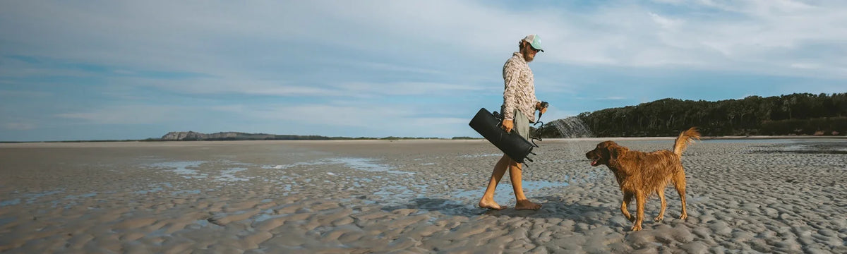 A man walks along a sandy beach with a dog, carrying a 4-gallon DayTank.