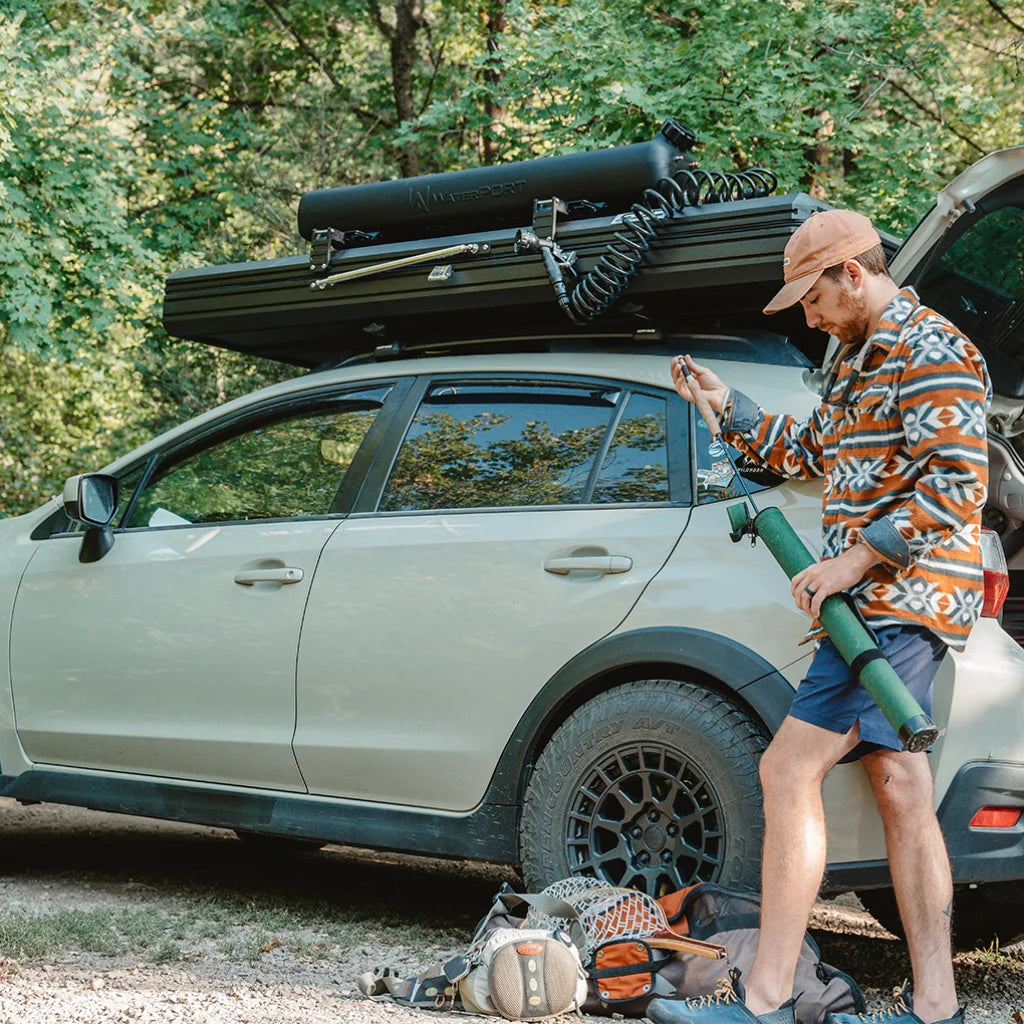 A man stands by a beige SUV with camping gear, including a fishing rod and backpack, preparing outdoors. The vehicle has equipment on the roof, and the background is green with trees.